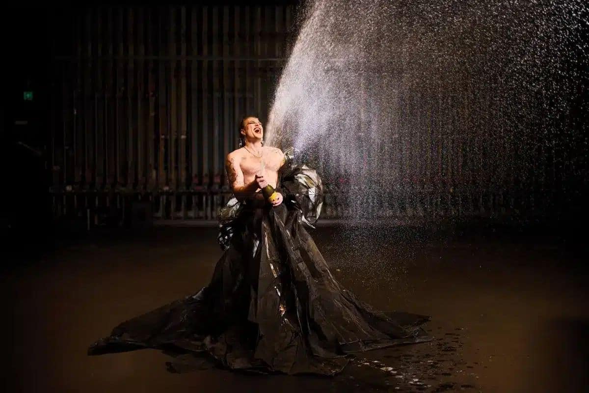 A shirtless performer in a dramatic black gown sprays champagne into the air inside a darkened industrial venue, laughing joyfully as mist cascades around them.