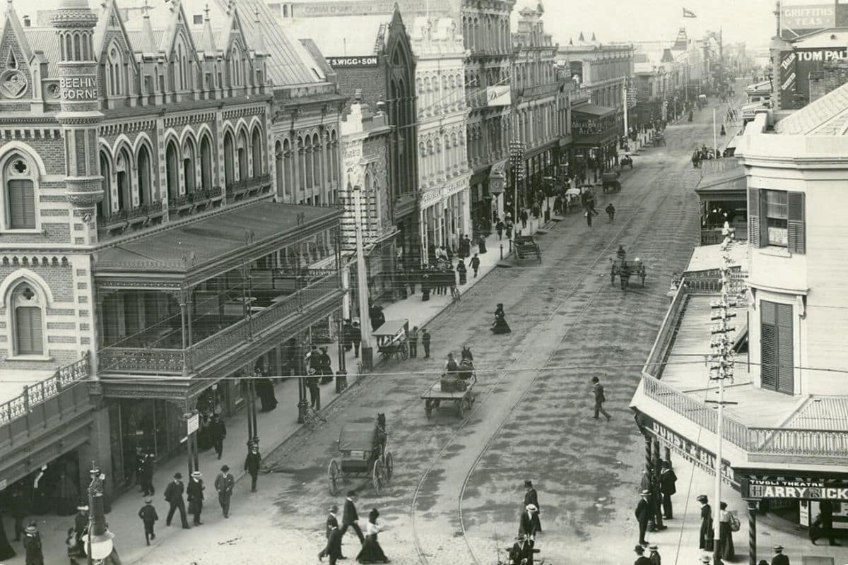 A black and white aerial photograph of Rundle Street in Adelaide's early colonial era, showing horse-drawn carriages, Victorian-era shopfronts including Beehive Corner, and pedestrians in period dress.
