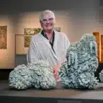 Artist Deb McKay stands smiling behind a display of large sculptural ceramic works resembling bleached coral formations, exhibited at the South Australian Museum.