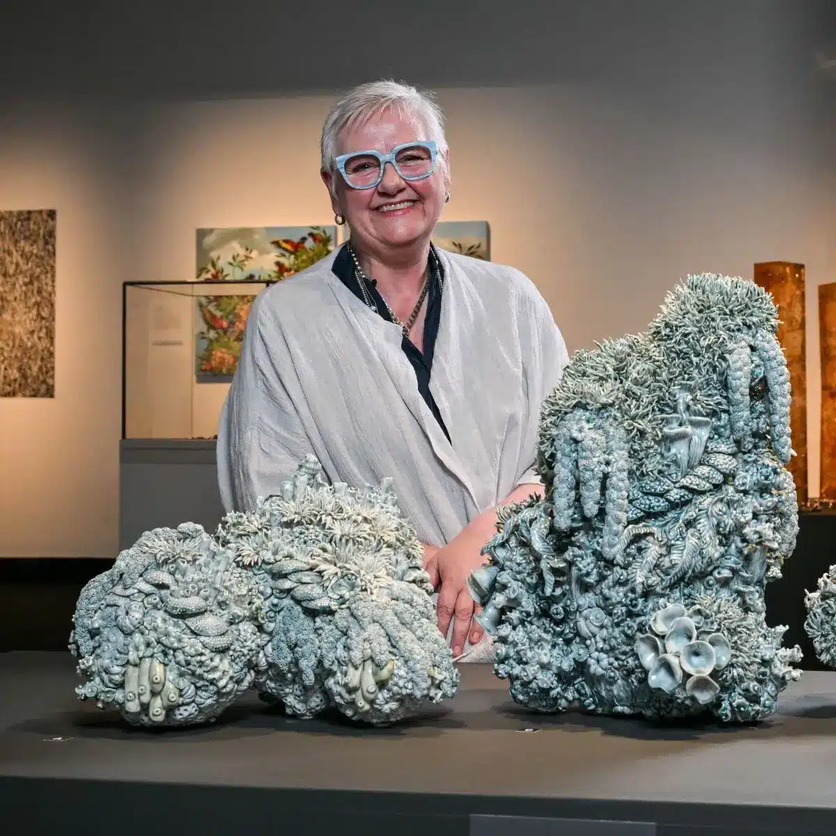 Artist Deb McKay stands smiling behind a display of large sculptural ceramic works resembling bleached coral formations, exhibited at the South Australian Museum.