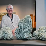 Artist Deb McKay stands smiling behind a display of large sculptural ceramic works resembling bleached coral formations, exhibited at the South Australian Museum.