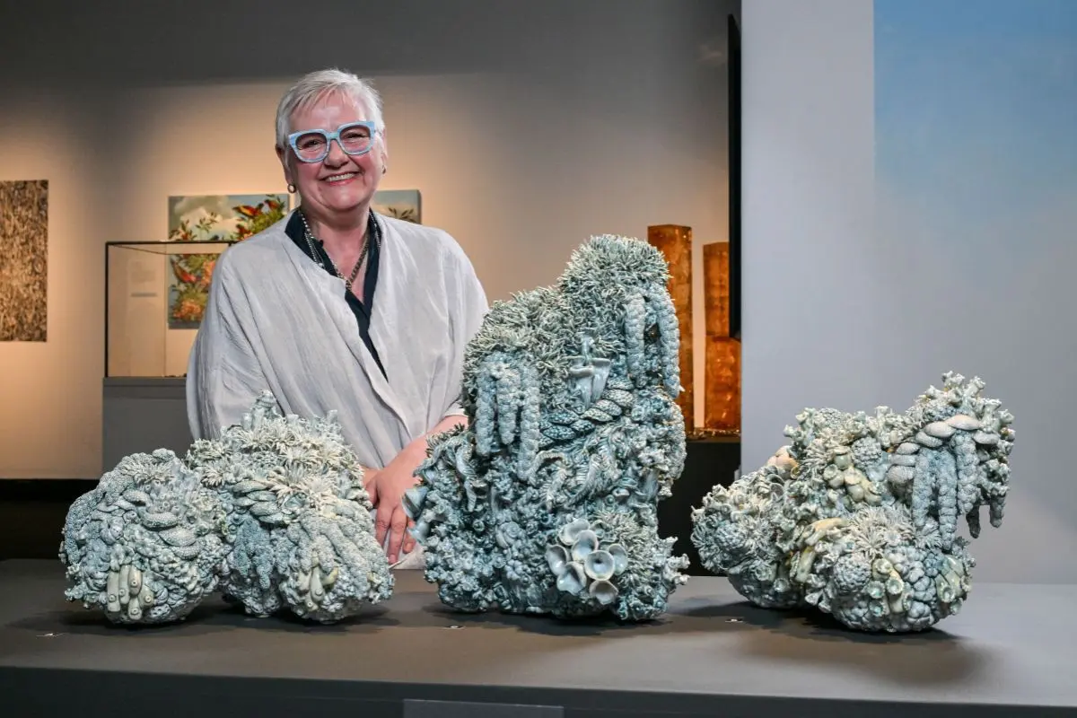 Artist Deb McKay stands smiling behind a display of large sculptural ceramic works resembling bleached coral formations, exhibited at the South Australian Museum.