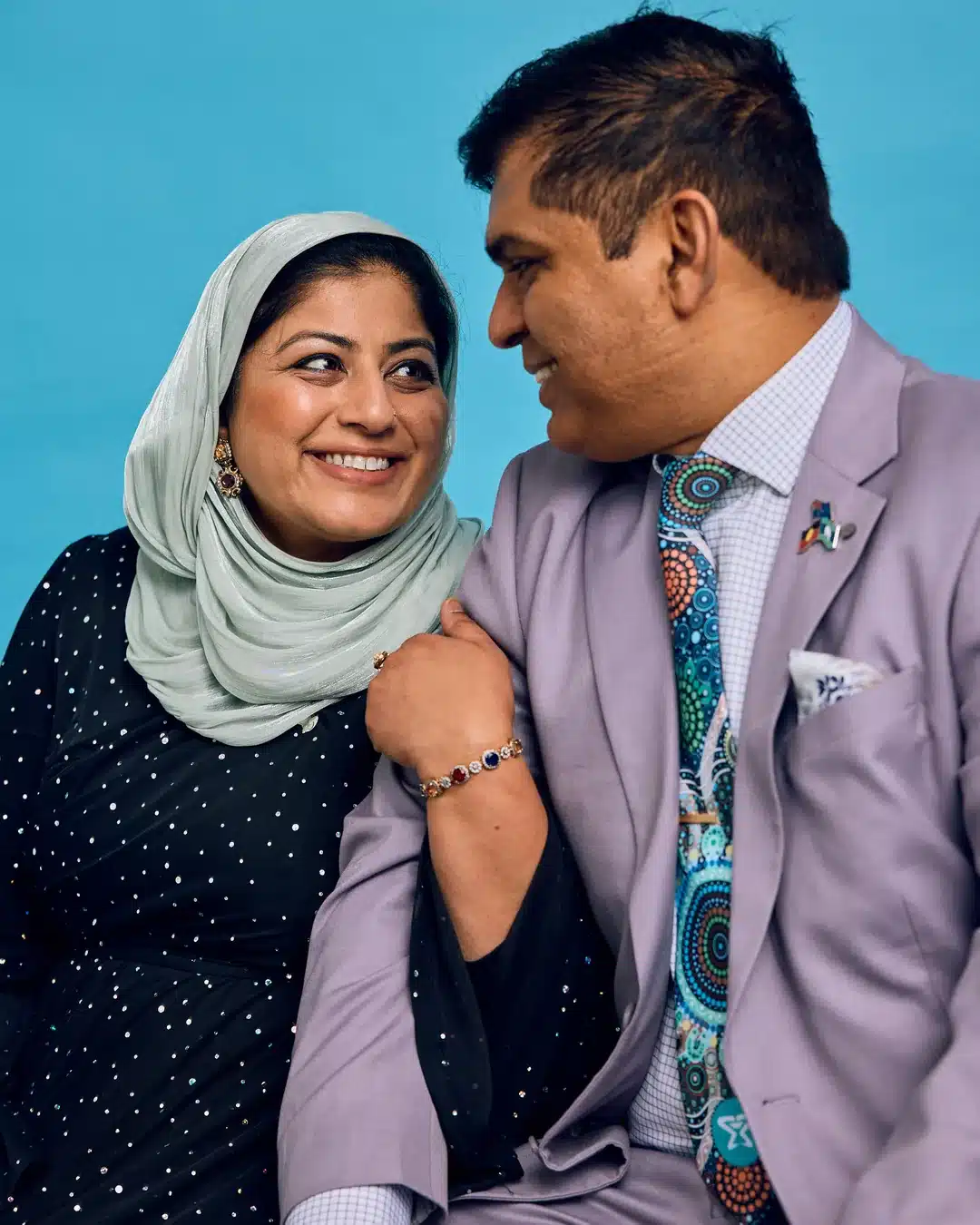 A couple smile warmly at each other against a bright teal studio backdrop. The woman wears a sage green hijab and a dark embellished dress; the man wears a lilac suit with a colourful Indigenous art-print tie.