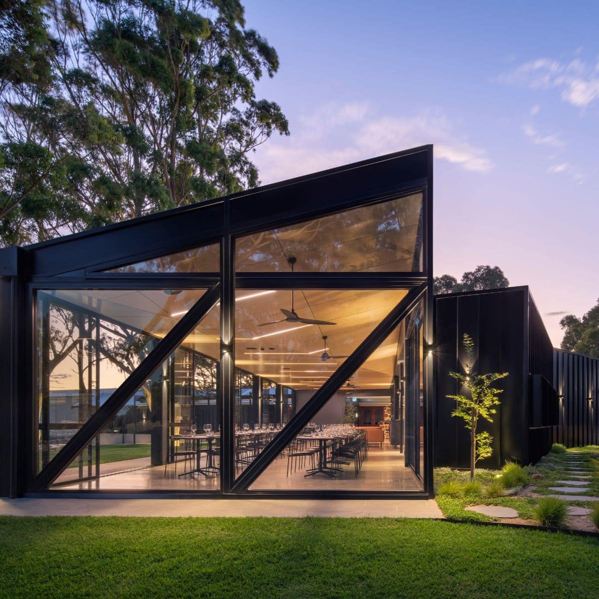 Modern glass-walled pavilion at Anvers Wines in the Adelaide Hills, set among greenery and trees, showcasing contemporary architecture and vineyard surroundings at dusk.