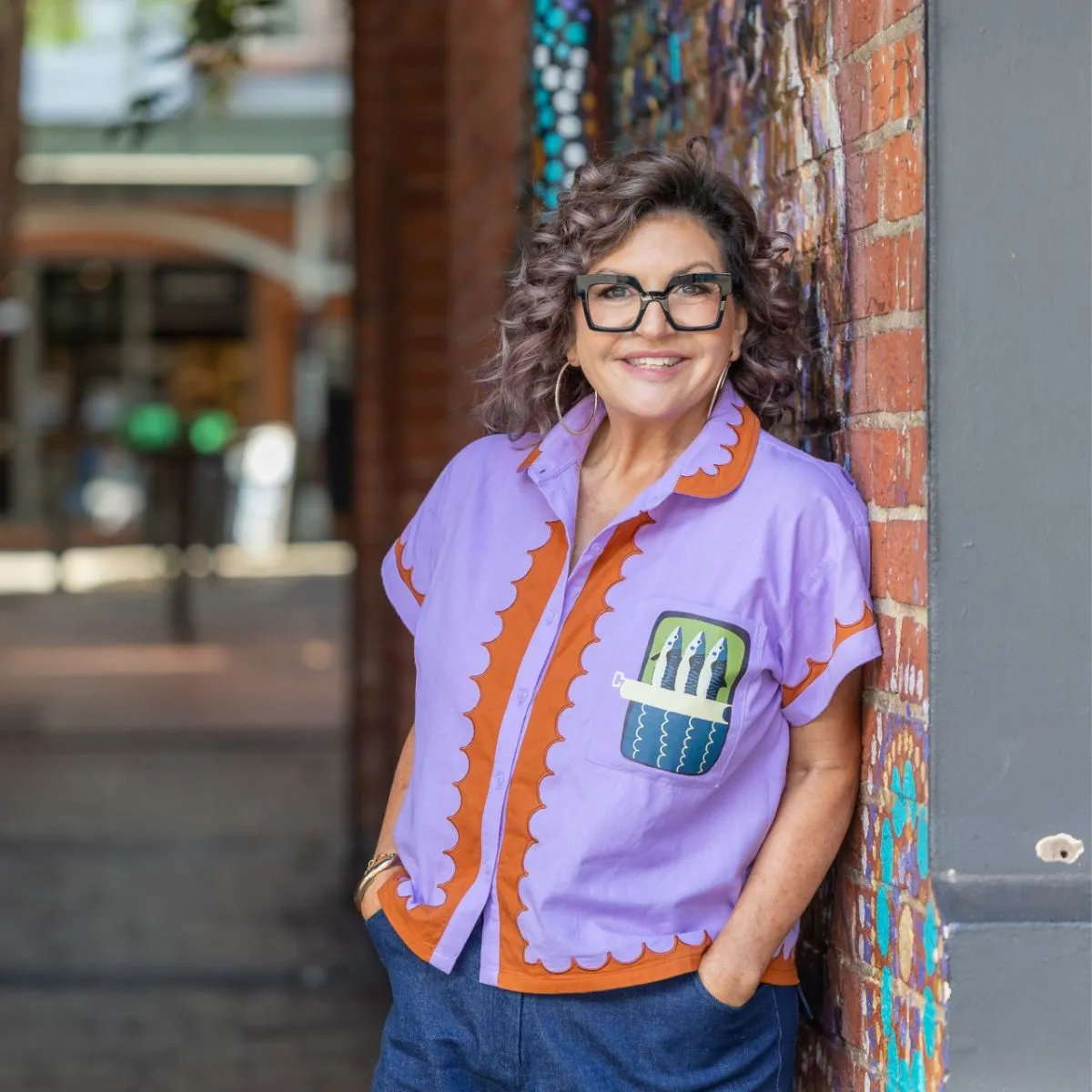 Mandy Hall standing against a wall in glasses and a vibrant purple shirt.