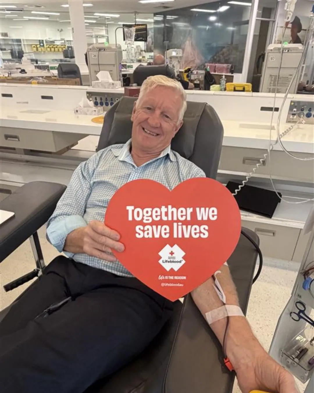 A man in a blue shirt sits in a chair while donating blood holing a loveheart signage.