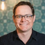 Adelaide Fringe CEO, Marc Carnes in black shirt and glasses against a green patterned background.