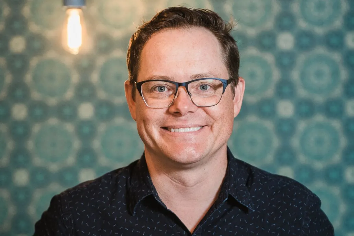 Adelaide Fringe CEO, Marc Carnes in black shirt and glasses against a green patterned background.