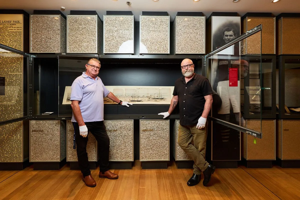 Two men wearing white conservation gloves stand either side of a glass display case housing the Hankey Panorama at the State Library of South Australia, surrounded by archival exhibition materials.