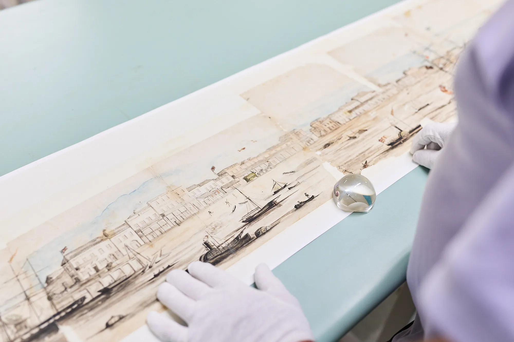 A conservator wearing white gloves carefully handles a long, aged watercolour panorama of Port Adelaide, by Frank George Hankey, laid flat on a light blue conservation table. The painting depicts a detailed colonial waterfront scene with sailing vessels, buildings and figures.