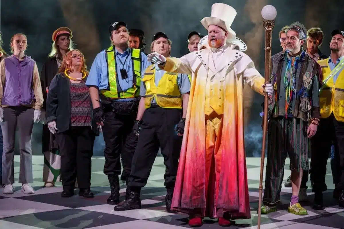 Teddy Tahu Rhodes in costume as Sarastro in a contemporary staging of The Magic Flute, commanding the stage in an ornate white robe and tall white hat, surrounded by ensemble cast members in high-visibility vests.