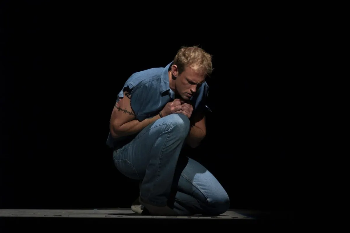
Teddy Tahu Rhodes crouches in a pool of light on a darkened stage, wearing a denim prison uniform, in a scene from Dead Man Walking.