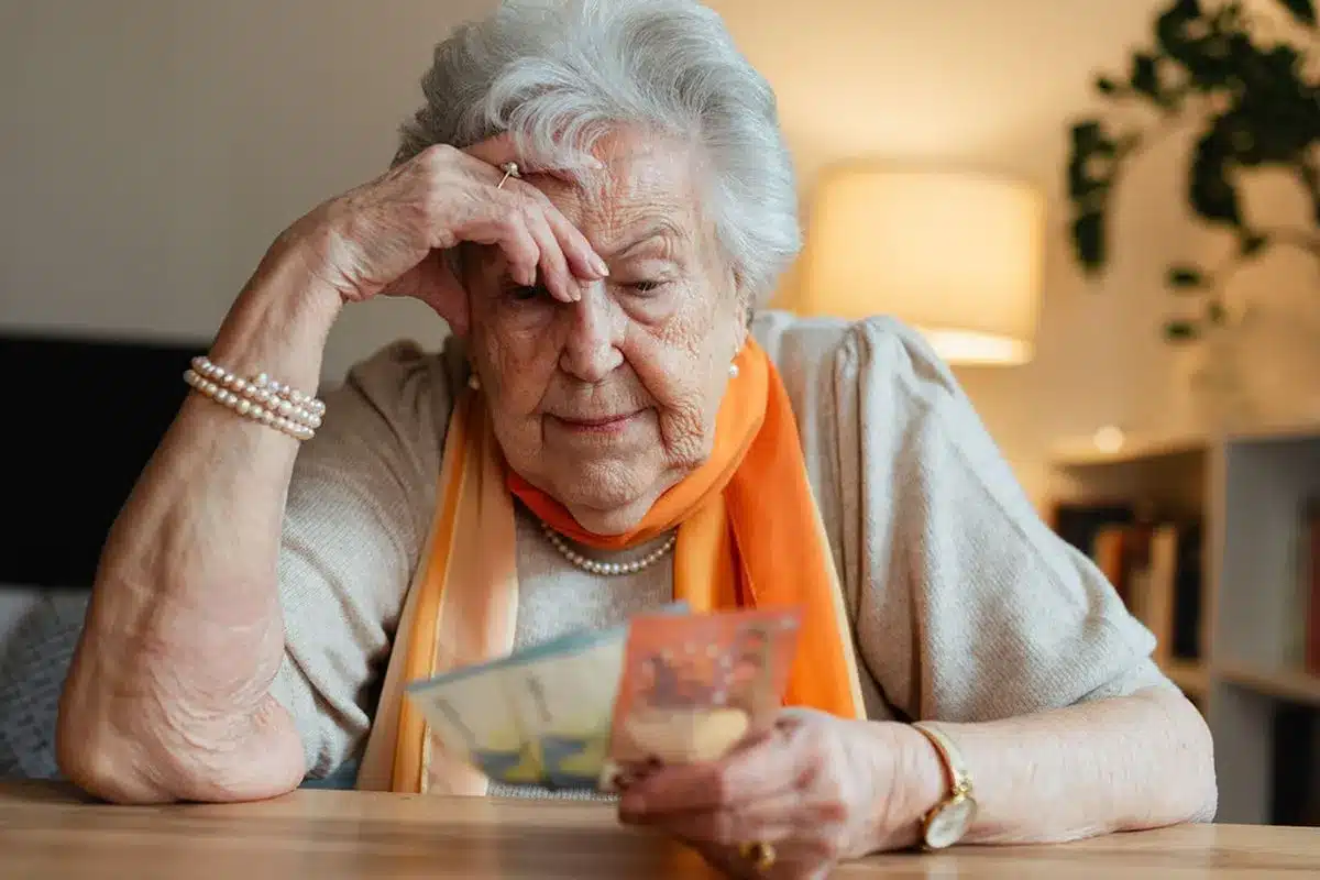 An older woman sits at a table at home, looking at bills with a concerned expression.