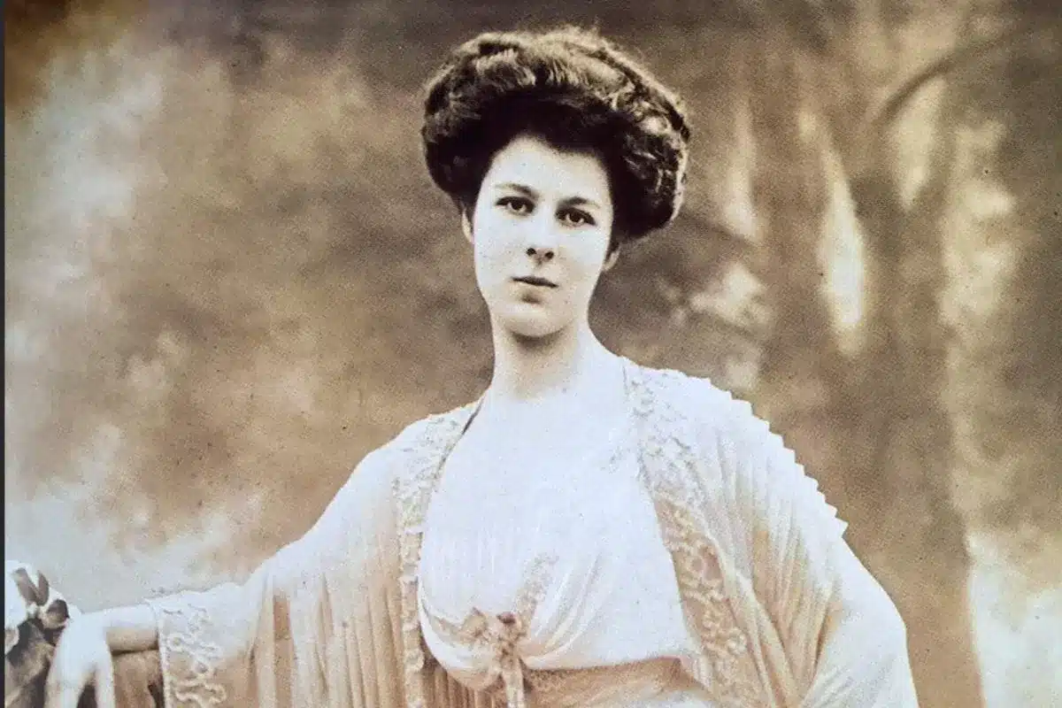 A sepia-toned Edwardian portrait photograph of a young woman in a delicate lace-trimmed gown, her dark hair pinned up in the style of the early 1900s.