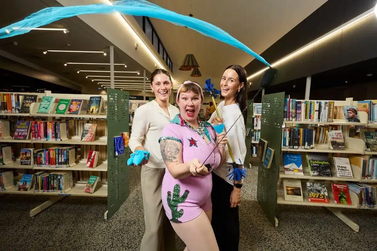 Erin Sundstrom, Lucy Frost and Kameron Bazzo performing circus tricks with a ribbon and cocktail glass among the shelves of a South Australian library for Firstival 2026
