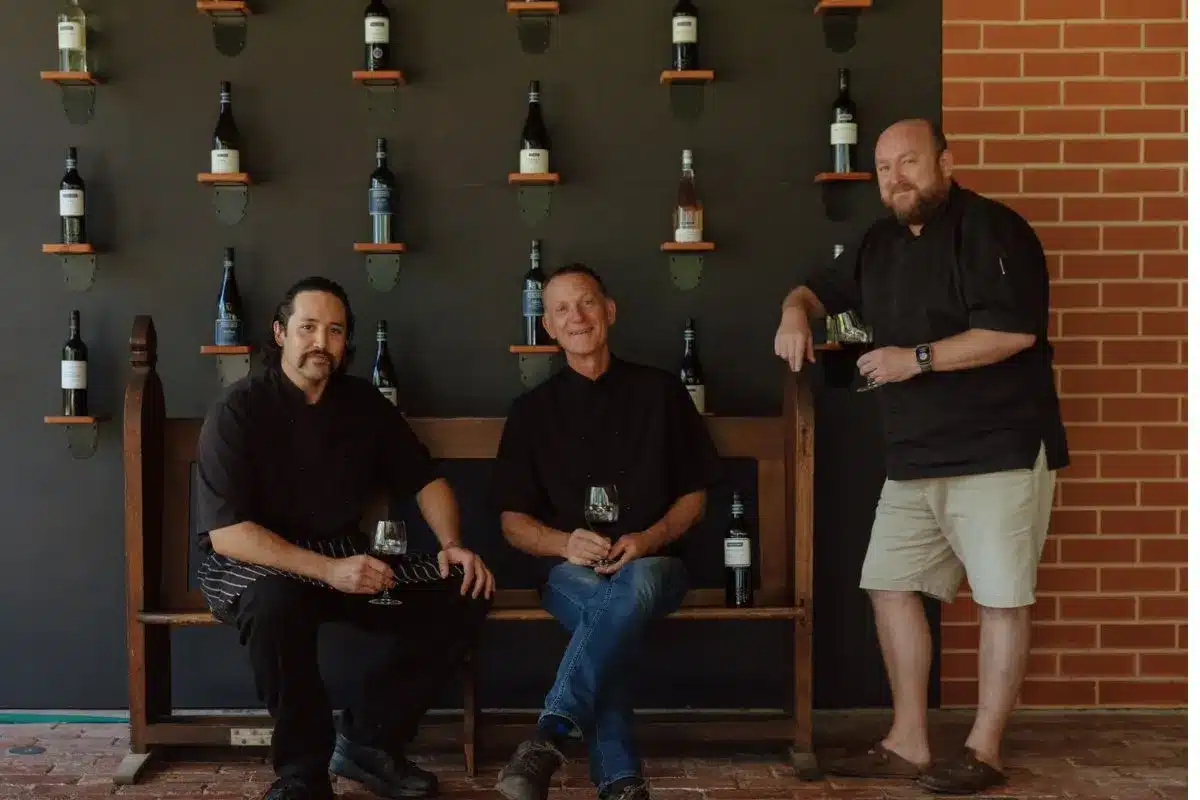 Head chef Taiaha Ngawiki and former Harry's Deli head chefs Rob Kolencik and Tom Boden seated together in the Wirra Wirra cellar door, with the winery's wine collection displayed on the wall behind them