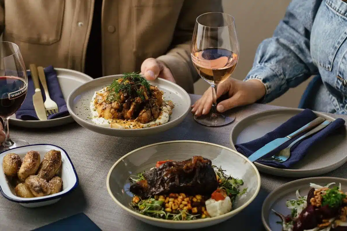 A table at Harry's Deli set with multiple winter menu dishes and Wirra Wirra wines, including a braised meat dish, mushroom risotto and roasted potatoes, with a glass of rosé being raised