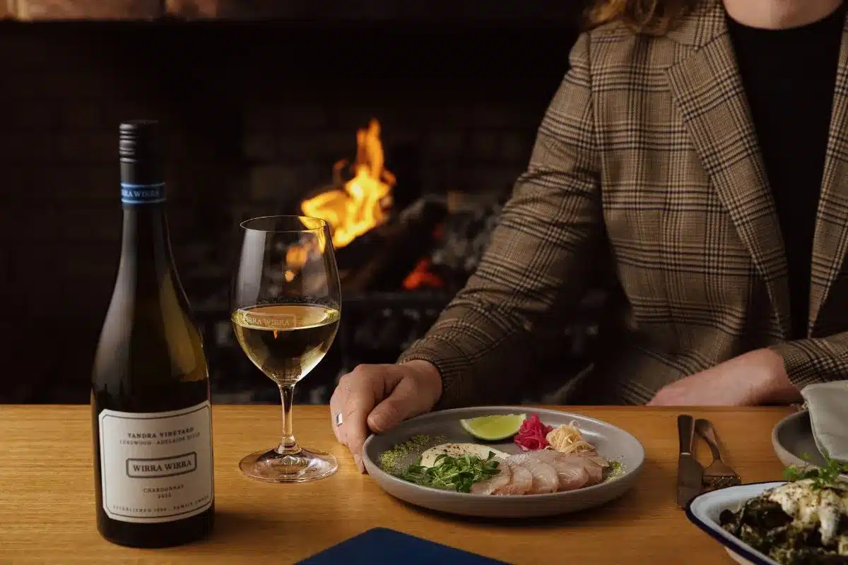 A diner reaches for a plate of kingfish sashimi beside a glass and bottle of Wirra Wirra Chardonnay, with an open fire burning in the background at Harry's Deli