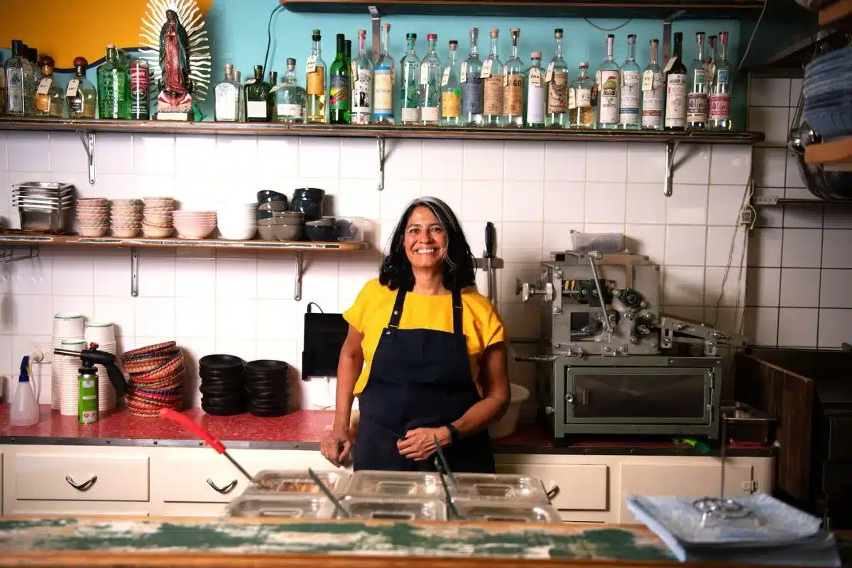Daniella Guevara Muñoz smiles behind the counter at La Popular Taqueria, with shelves of mezcal and tequila bottles lining the wall behind her and the restaurant's tortilla press visible to the right