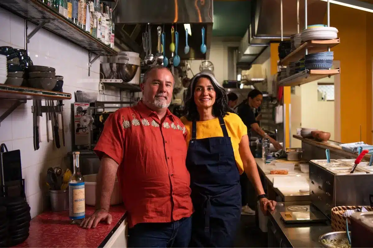 Daniella Guevara Muñoz and Kor-jent van Dijk stand together in the kitchen of La Popular Taqueria in Port Adelaide, smiling for the camera amid shelves of cookware and hanging utensils