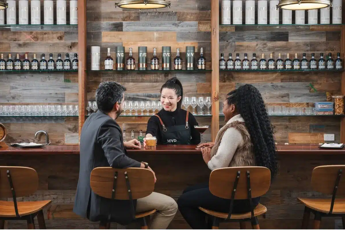 a couple sit at a cellar door bar counter served by a waiter in an apron.