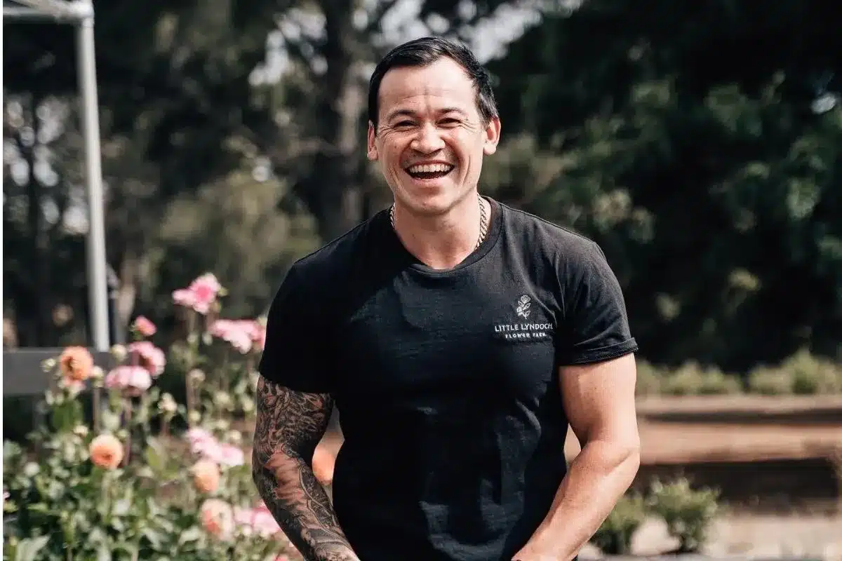 A smiling person wearing a Little Lyndoch Flower Farm t-shirt stands among pink and peach blooms at the Barossa Valley flower farm