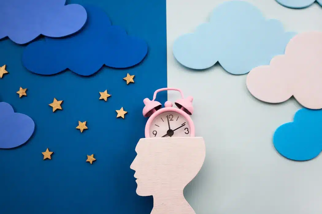 A wooden silhouette of a human head with a pink alarm clock emerging from the top, set against a blue background with gold stars, representing sleep and the mind.