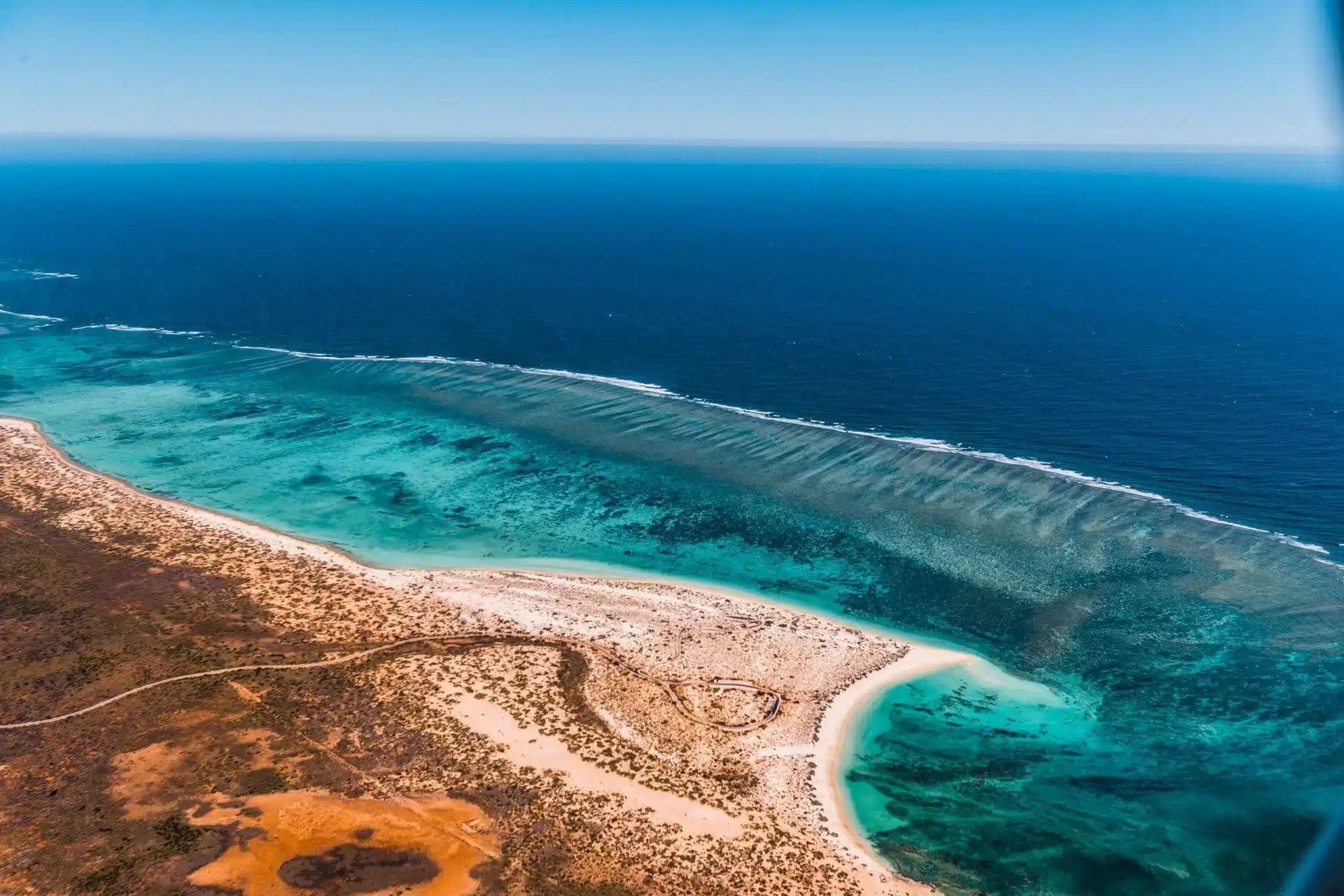 A high aerial view of the Ningaloo Reef coastline near Exmouth, showing the full stretch of the fringing reef where red desert scrubland meets the deep blue Indian Ocean
