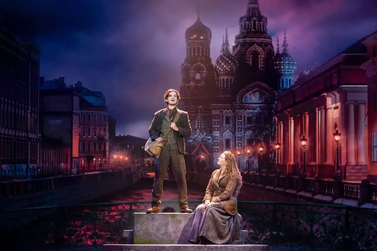 Two actors on stage as Anya and Dmitry in ANASTASIA, standing on a bridge with an illuminated Russian cathedral backdrop