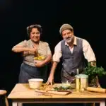 Alexis West and Ali Ammouchi in costume for Logan St, standing behind a wooden table set with bread, herbs and cooking ingredients.