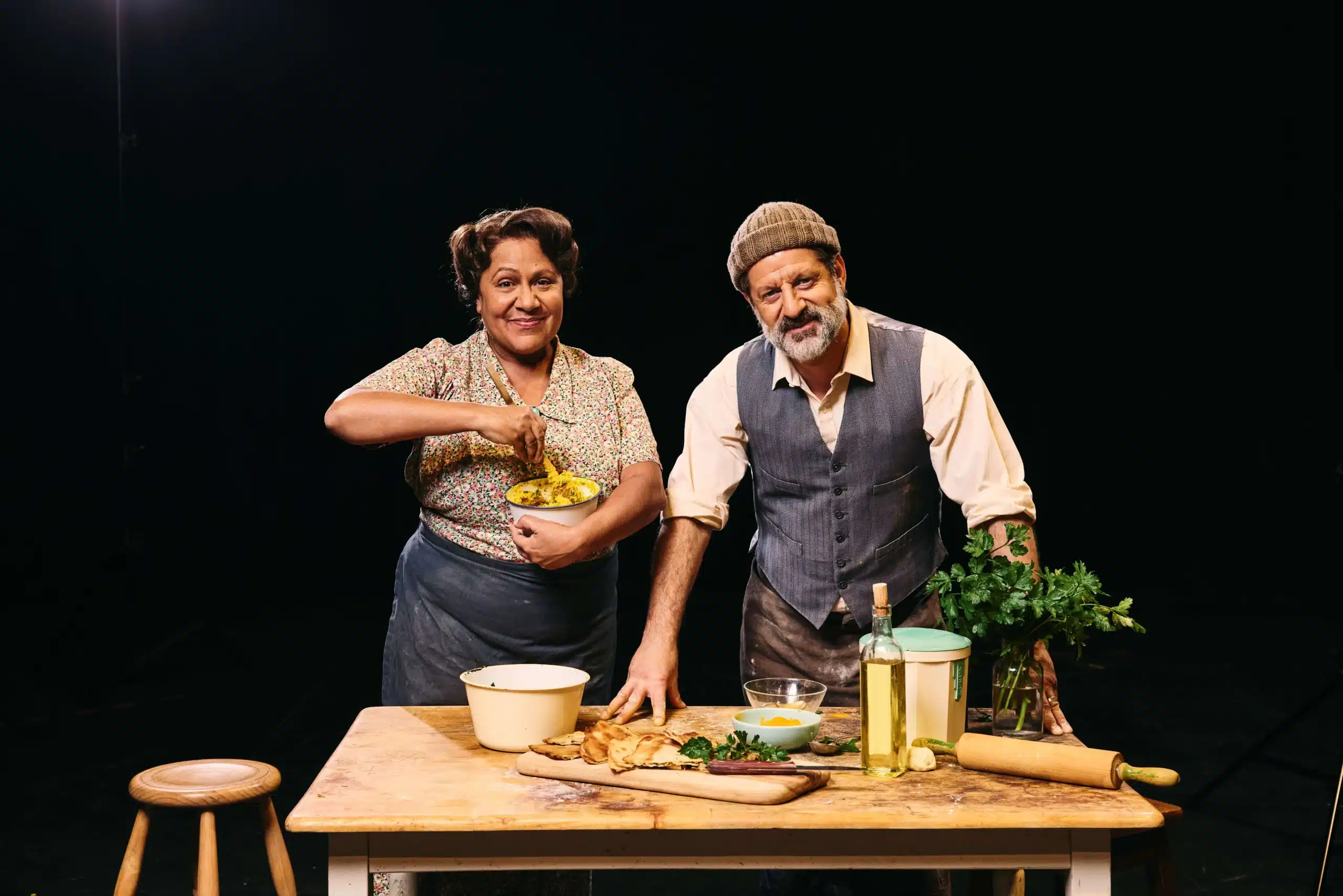 Alexis West and Ali Ammouchi in costume for Logan St, standing behind a wooden table set with bread, herbs and cooking ingredients.