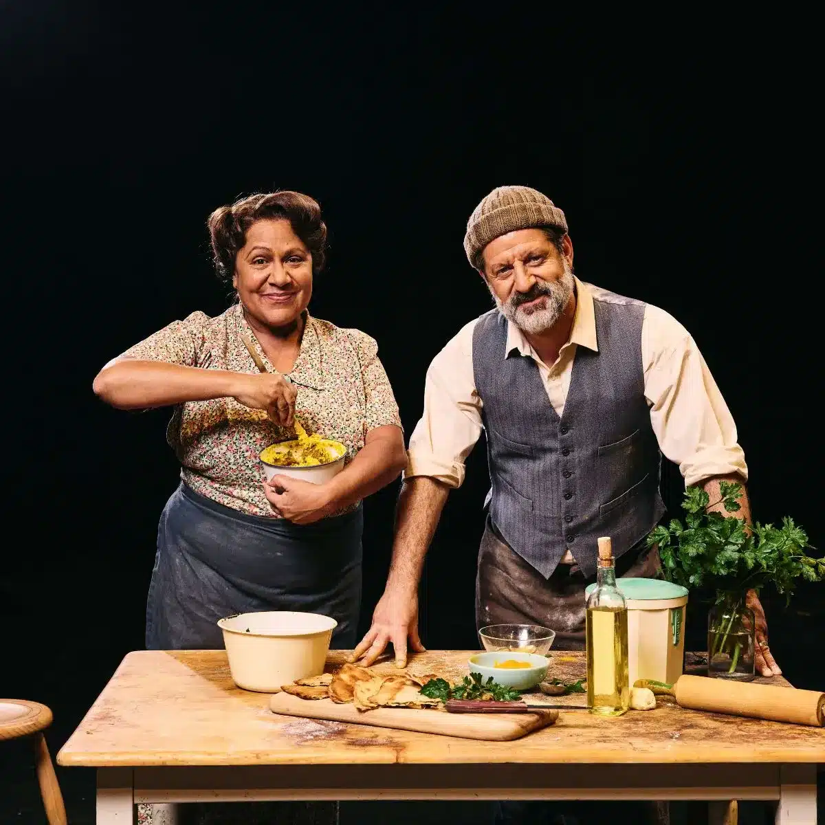 Alexis West and Ali Ammouchi in costume for Logan St, standing behind a wooden table set with bread, herbs and cooking ingredients.