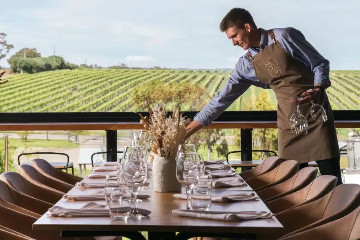 Waiter setting a long lunch table overlooking vineyard rows at Maxwell Wines, McLaren Vale.