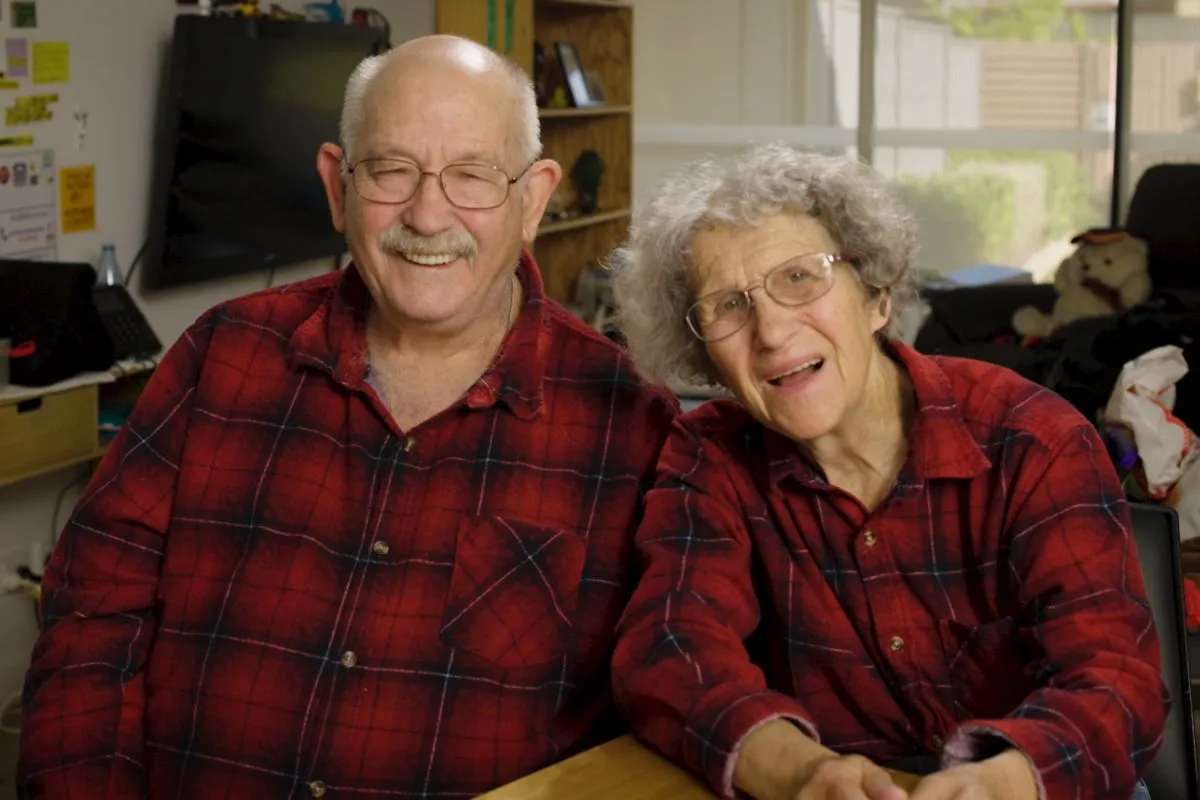 Two people featured in Minda's documentary A House for Humans, sitting together and smiling in matching red plaid shirts.