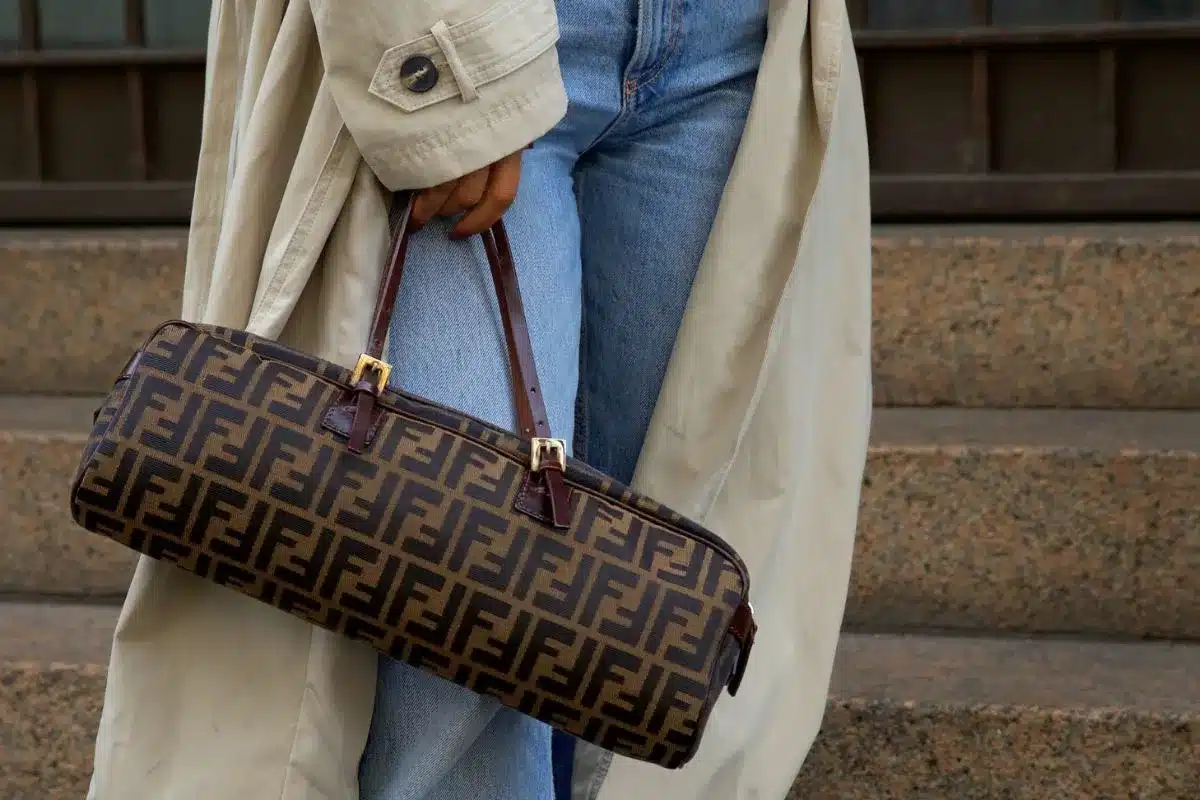 Woman in a beige trench coat and denim jeans carrying a brown Fendi Zucca monogram handbag on stone steps.