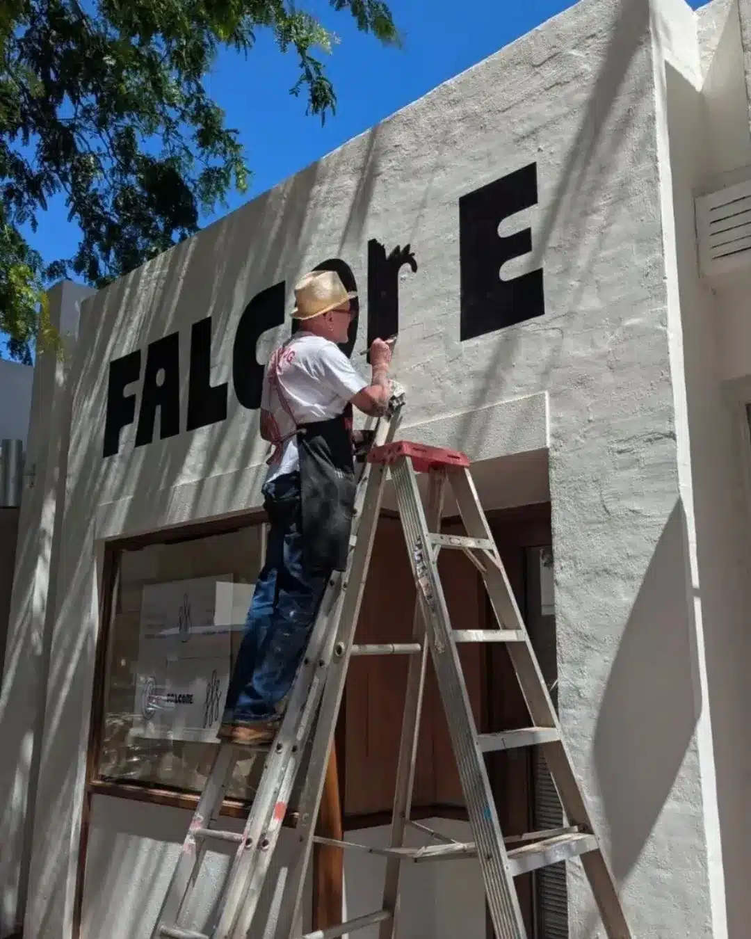 Raymond P MoonDog hand-painting large black lettering on a shopfront wall while standing on a ladder.