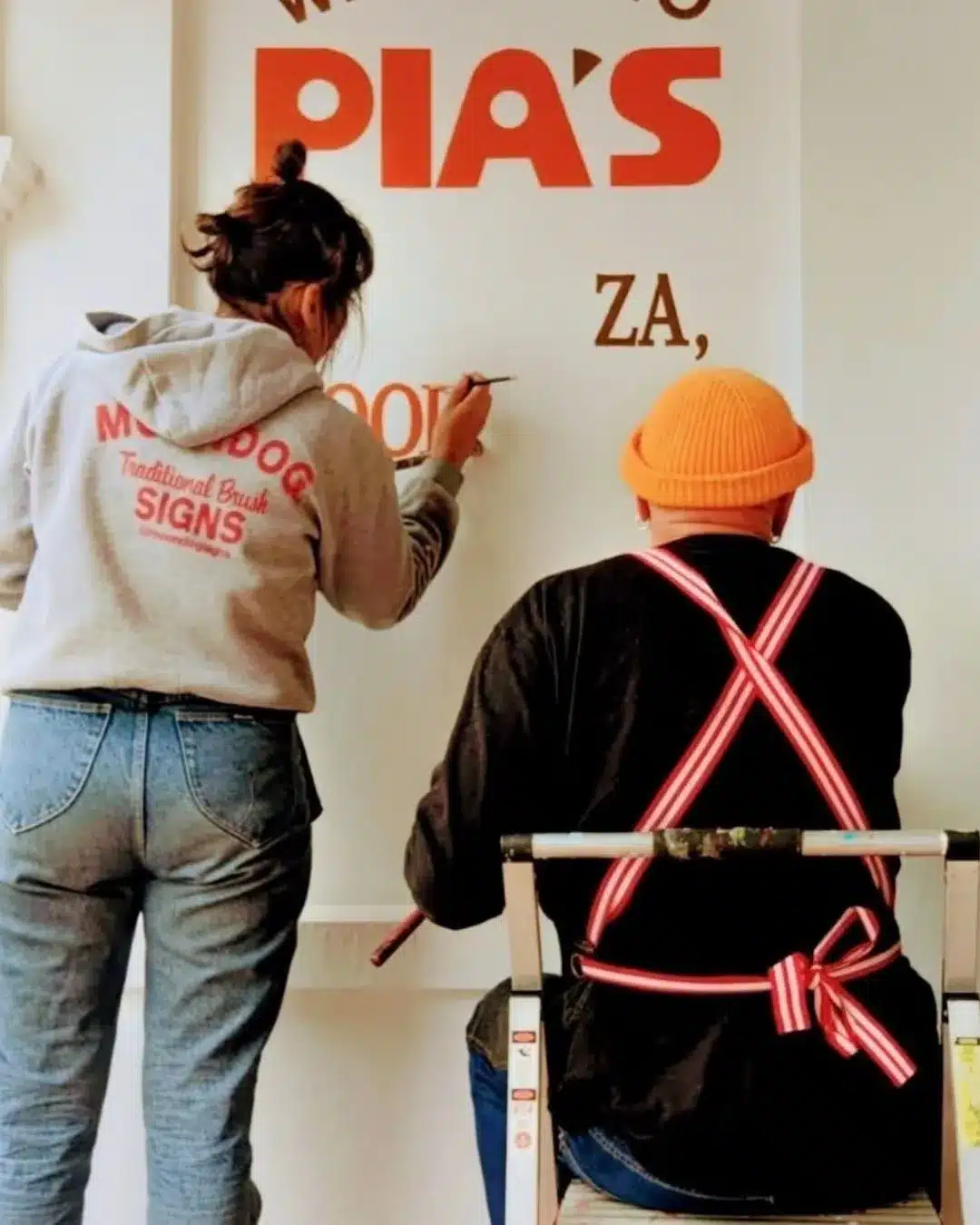 Raymond P MoonDog and an assistant hand-painting red PIA'S signage on a shopfront wall.
