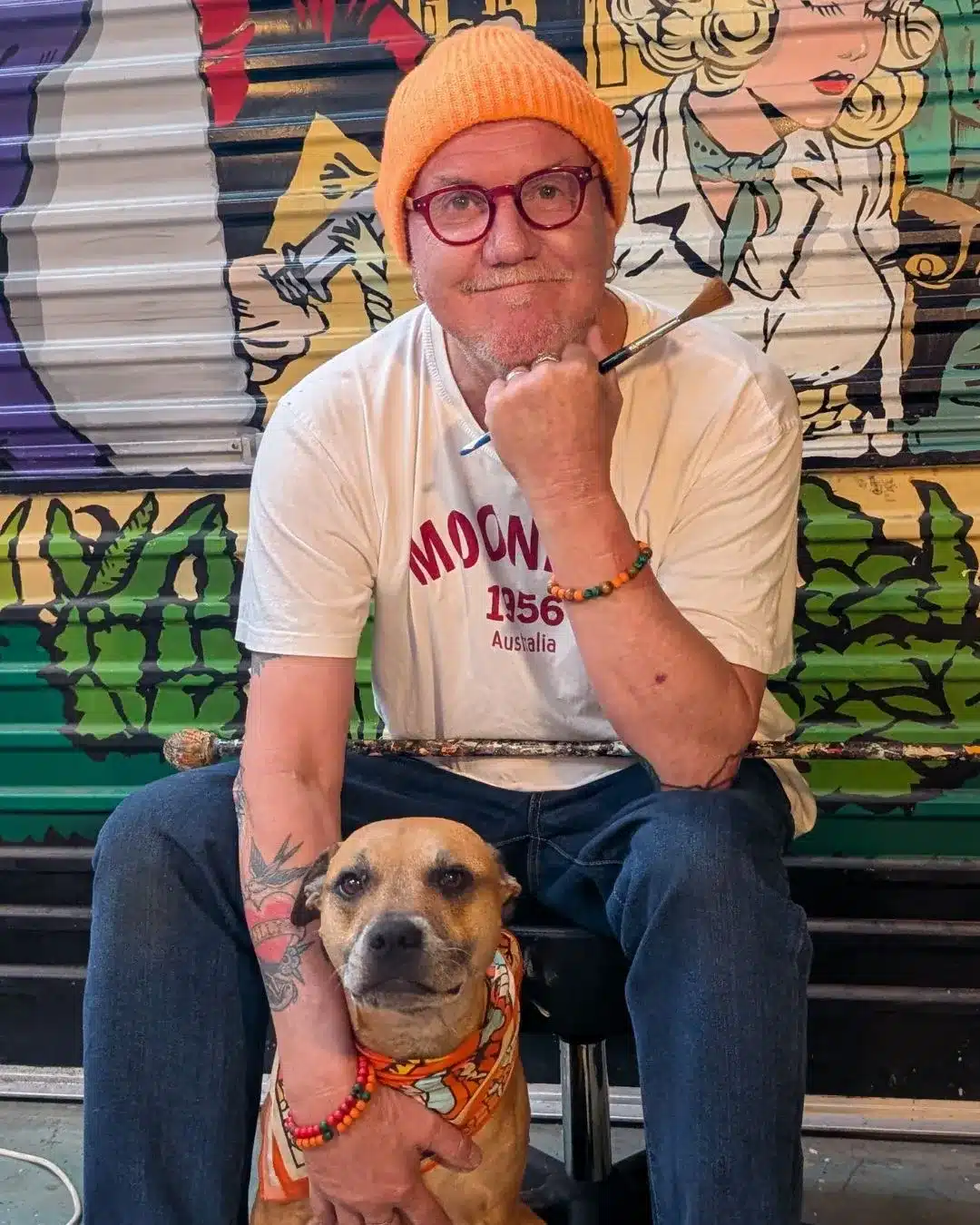 Signwriter Raymond P MoonDog seated in front of a colourful mural wearing a yellow beanie, with a dog beside him.