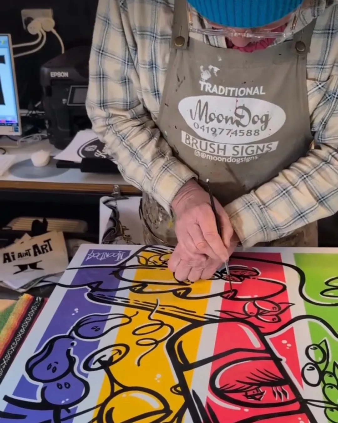 Raymond P MoonDog hand-painting colourful line art in his studio, wearing a MoonDog Traditional Brush Signs apron.