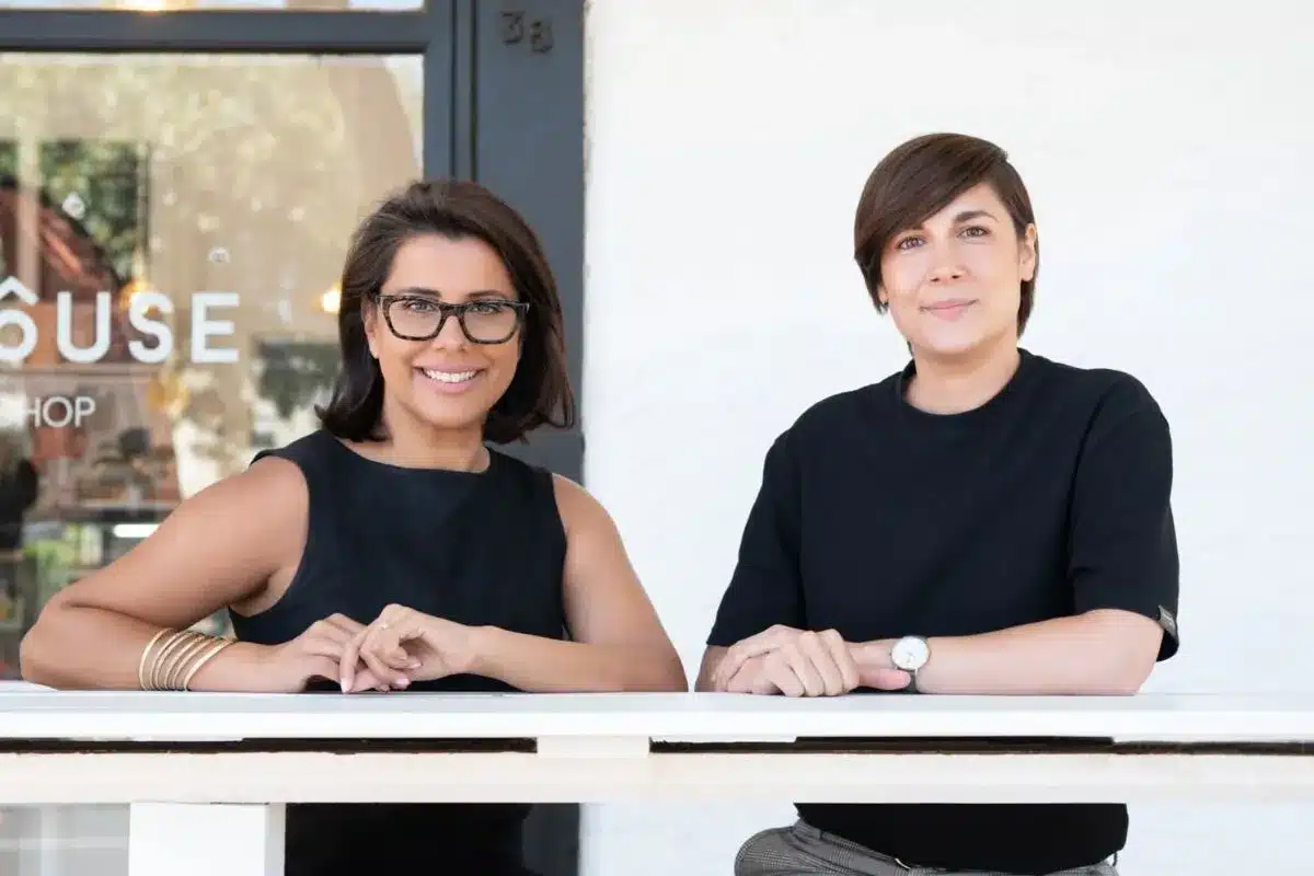 Two women wearing black tops stand side by side, leaning on a white railing outside a storefront.