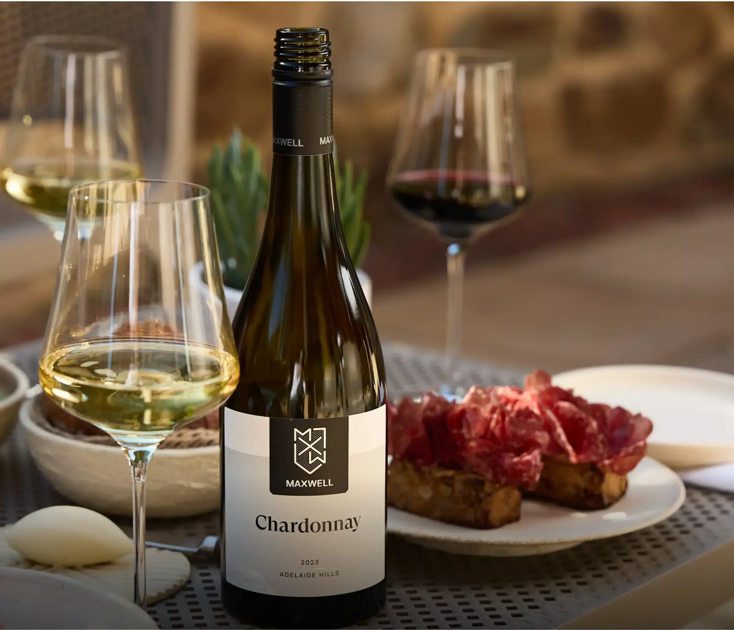 Bottle of Maxwell Chardonnay 2023 surrounded by glasses of wine and shared food on a table.