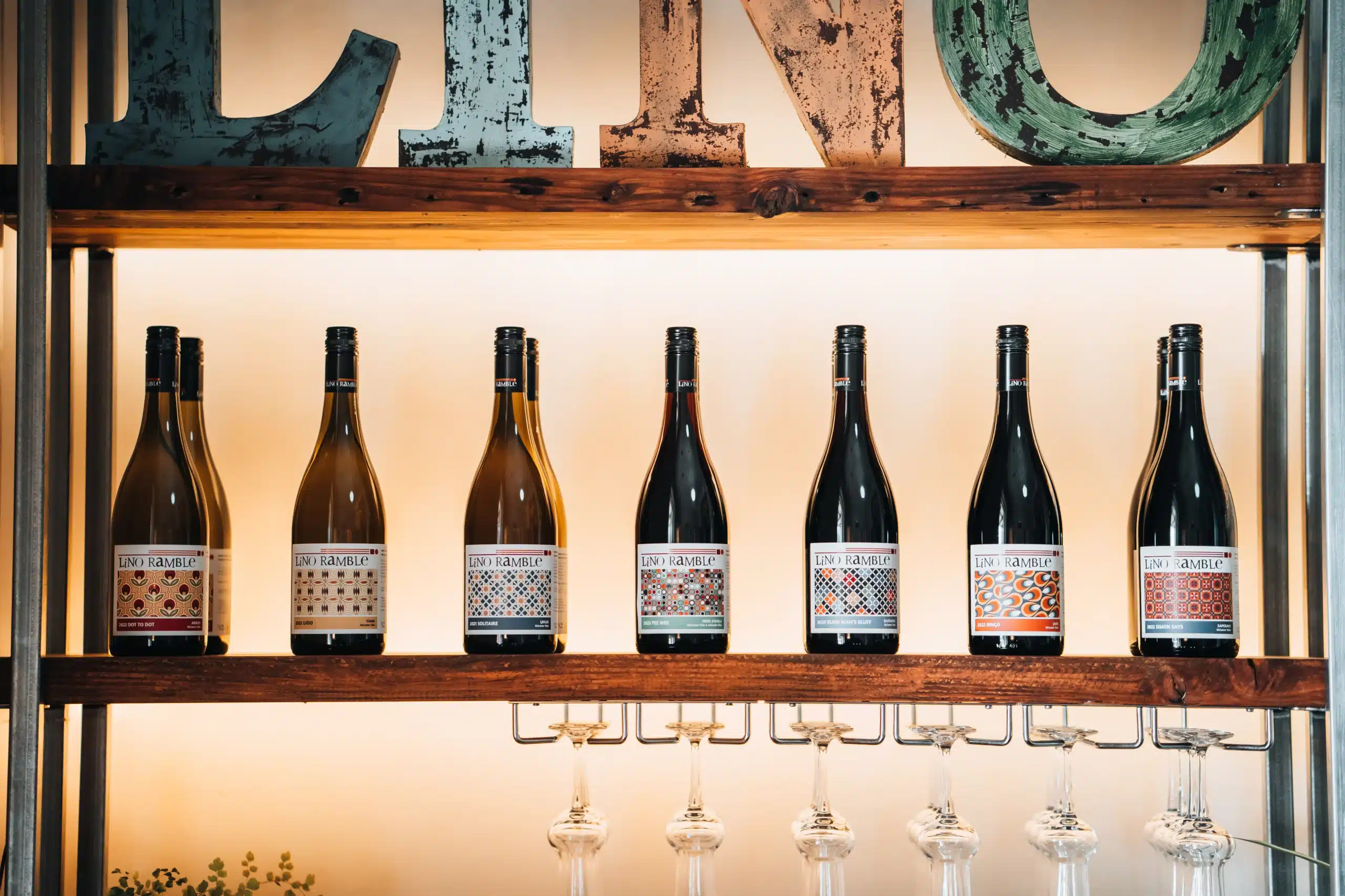 Row of Lino Ramble wine bottles on a shelf beneath a rustic "Lino" sign, with glasses hanging below.